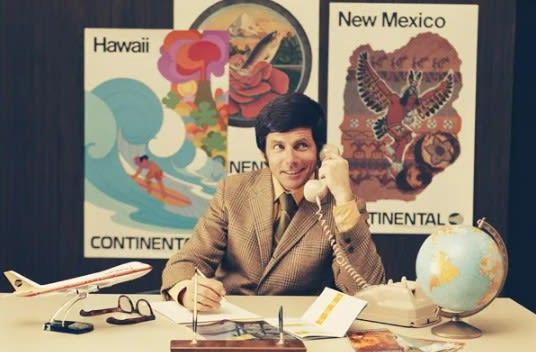 A man sitting at a desk full of travel knick-knacks, on the phone